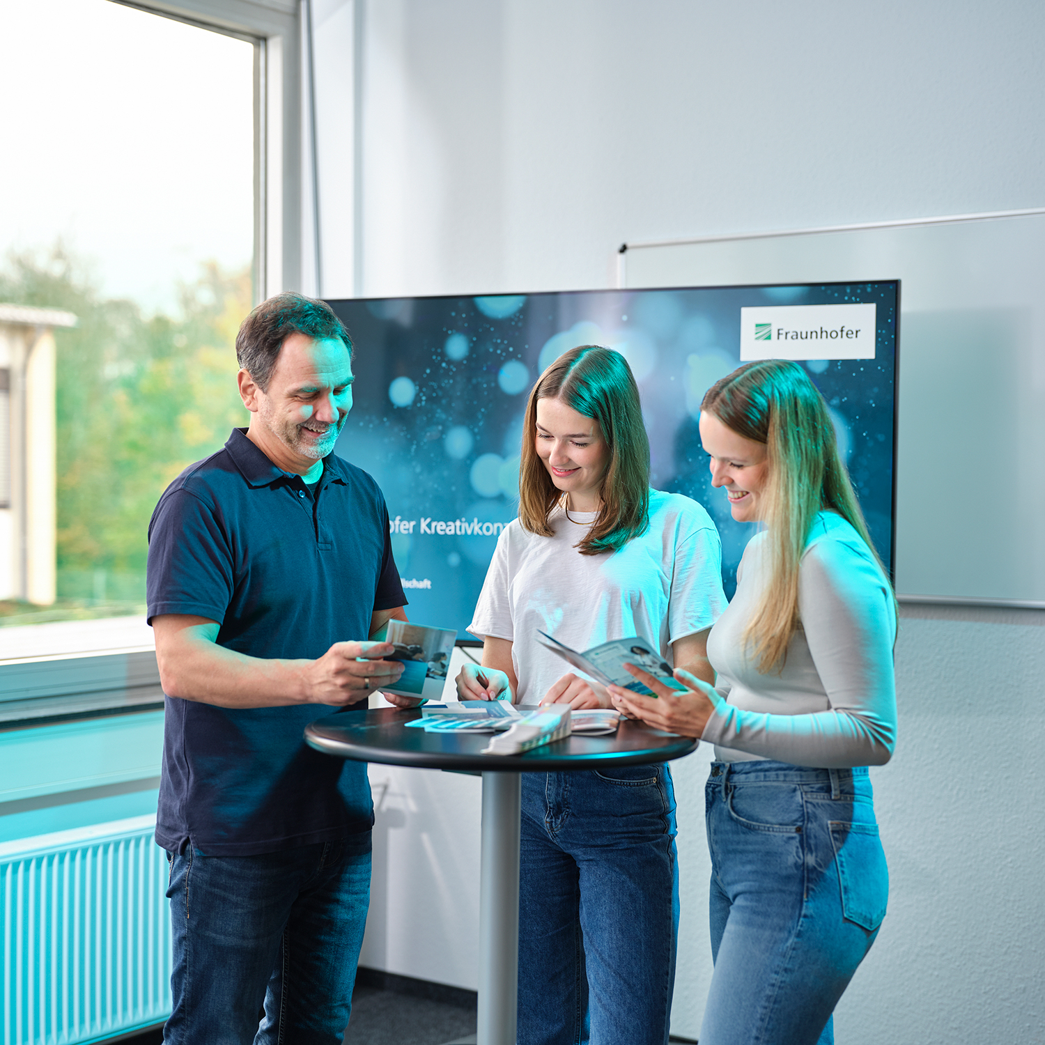 Ein Mann und zwei Frauen, eine mit Tablet in der Hand, sitzen an einem Meeting-Tisch. Eine weitere Frau steht hinter ihnen.