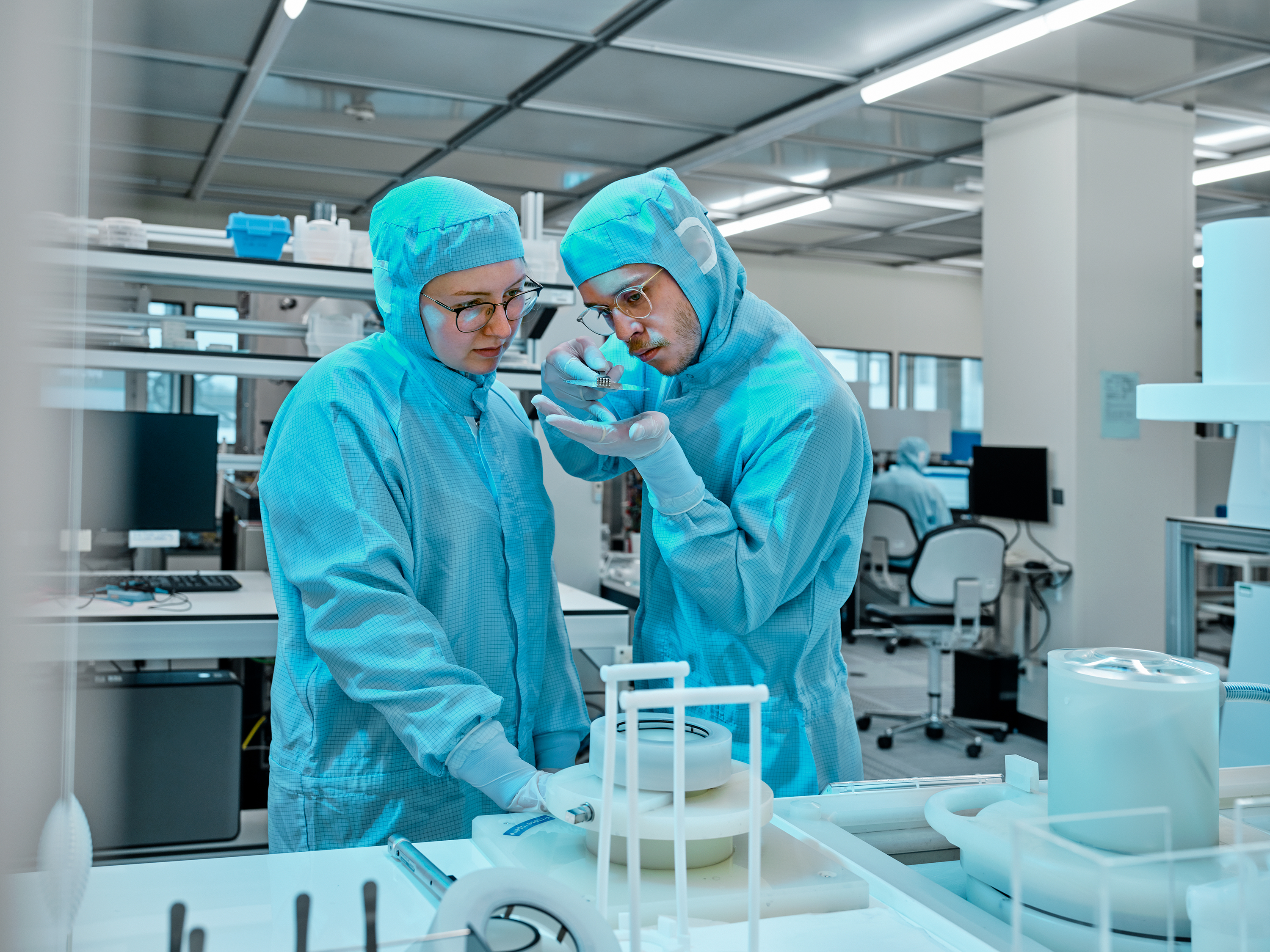 Eine Frau und ein Mann in Schutzanzügen stehen in einem Labor und schauen konzentriert auf einen kleinen Gegenstand in der Hand des Mannes.