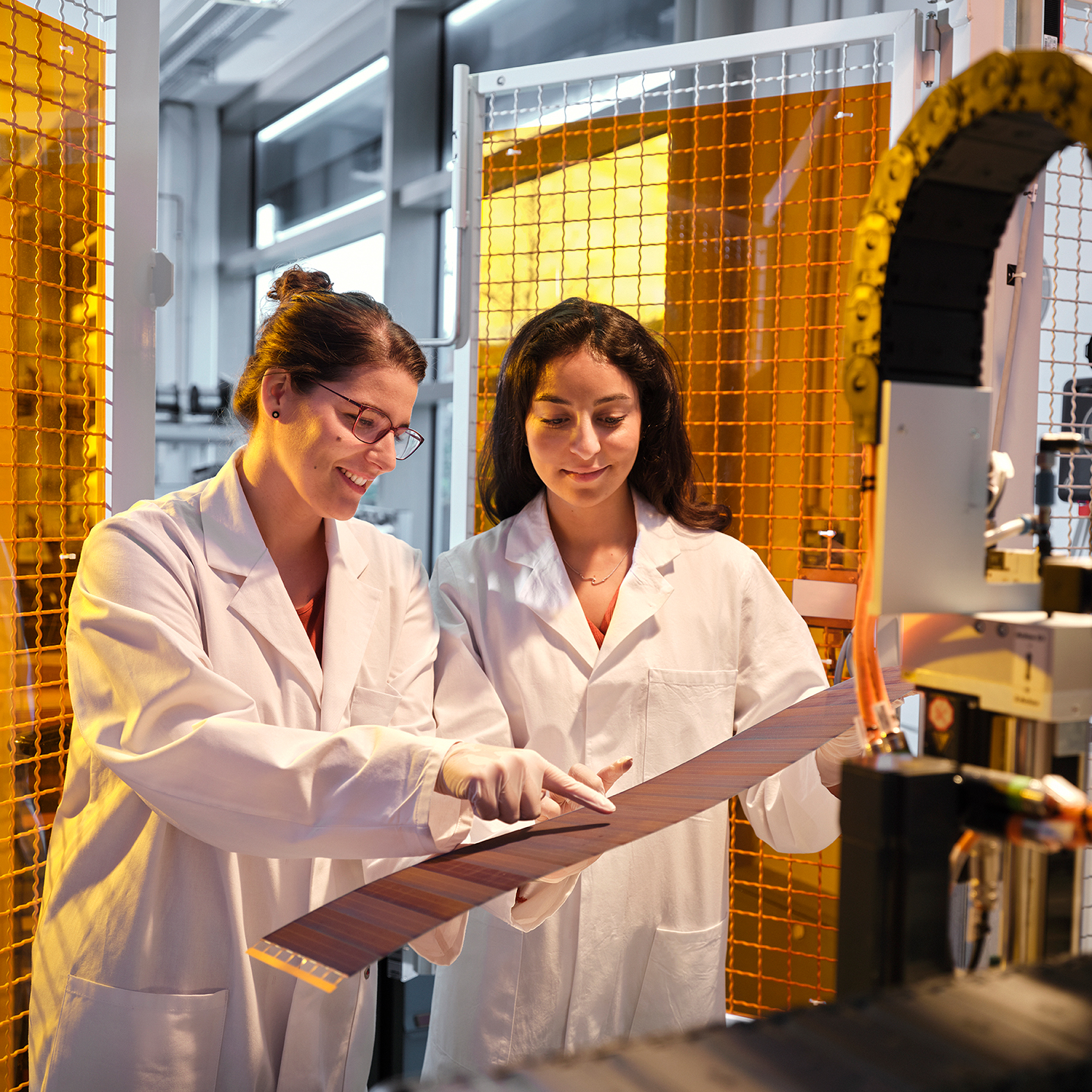 Zwei Frauen in Laborkitteln stehen vor einem Tisch mit technischen Geräten. Eine Frau hält eine Platte in der Hand.